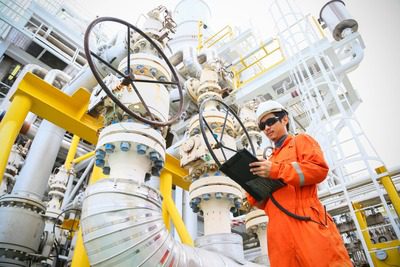 A worker in an orange safety uniform and helmet is inspecting industrial equipment at an oil refinery or chemical plant. They are holding a tablet, surrounded by large pipes, valves, and metal structures. The setting is outdoors, with complex machinery and infrastructure visible in the background.