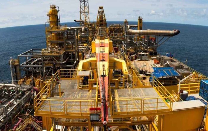 An offshore oil platform with intricate yellow structures, pipelines, and machinery, set against the backdrop of a vast ocean under a clear blue sky.