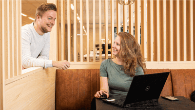 Woman at laptop smiling and talking to man in modern office setting.