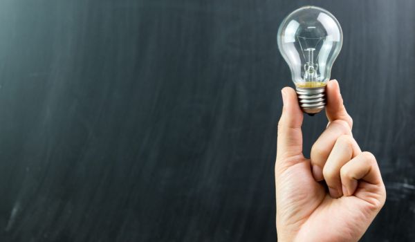 The image shows a hand holding a light bulb delicately by the base, set against a dark background.