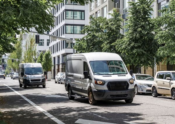 Two gray delivery vans on city street with trees and modern buildings.