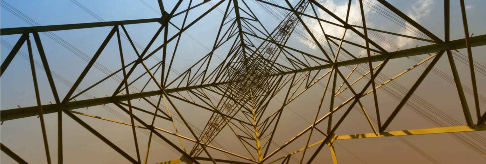 Upward view of steel tower beams and power lines against blue and orange sky.