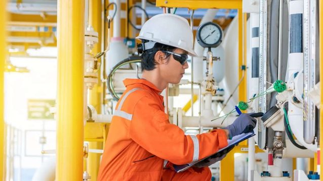 Engineer in protective gear inspecting industrial equipment with a clipboard.