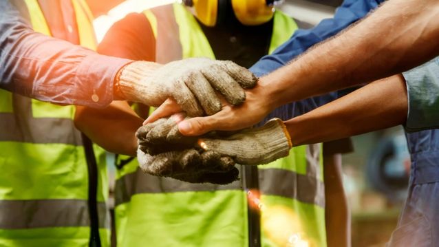 Workers wearing safety vests and gloves stacking their hands together in a teamwork gesture.