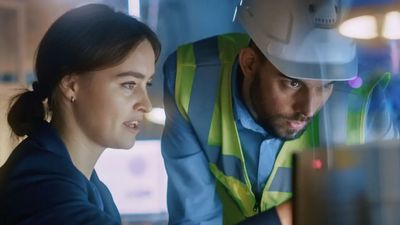 A woman in business attire and a man in a hard hat and high-visibility vest collaborate in a high-tech industrial environment, analyzing data on a computer screen.